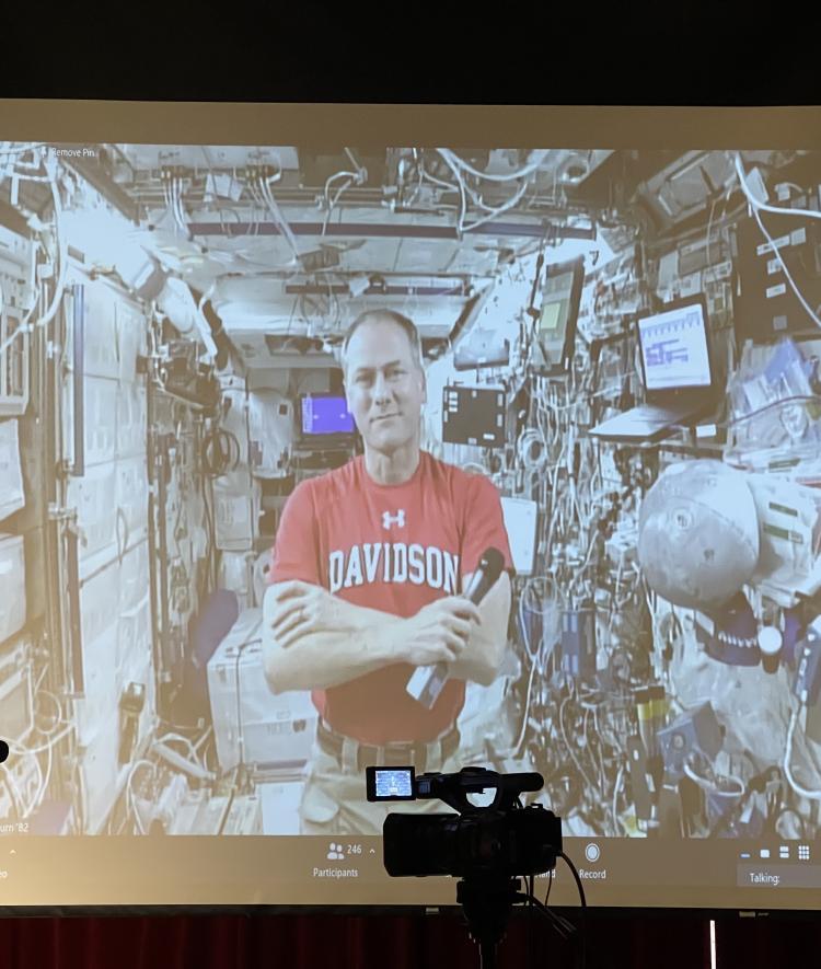 Tom Marshburn in a  t-shirt on a spaceship shown on a projector screen