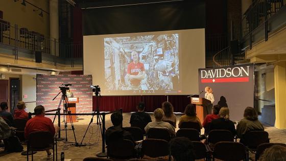 students seated in the 900 room, looking toward the front at Tom Marshburn on a screen in a  t-shirt in a spaceship with two in-person people behind podiums on both sides of the screen