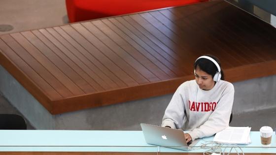 student wearing a  sweatshirt and headphones works on a laptop in a modern academic building