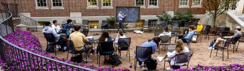  College Professor Hammurabi Mendes teaches class students outdoors with chalkboard surrounded by purple flower bushes