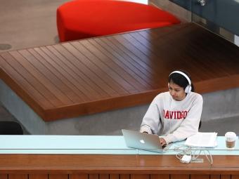 student wearing a  sweatshirt and headphones works on a laptop in a modern academic building