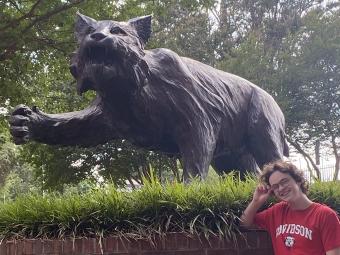a young student wears a  t-shirt while smiling and standing in front of a wildcat statue